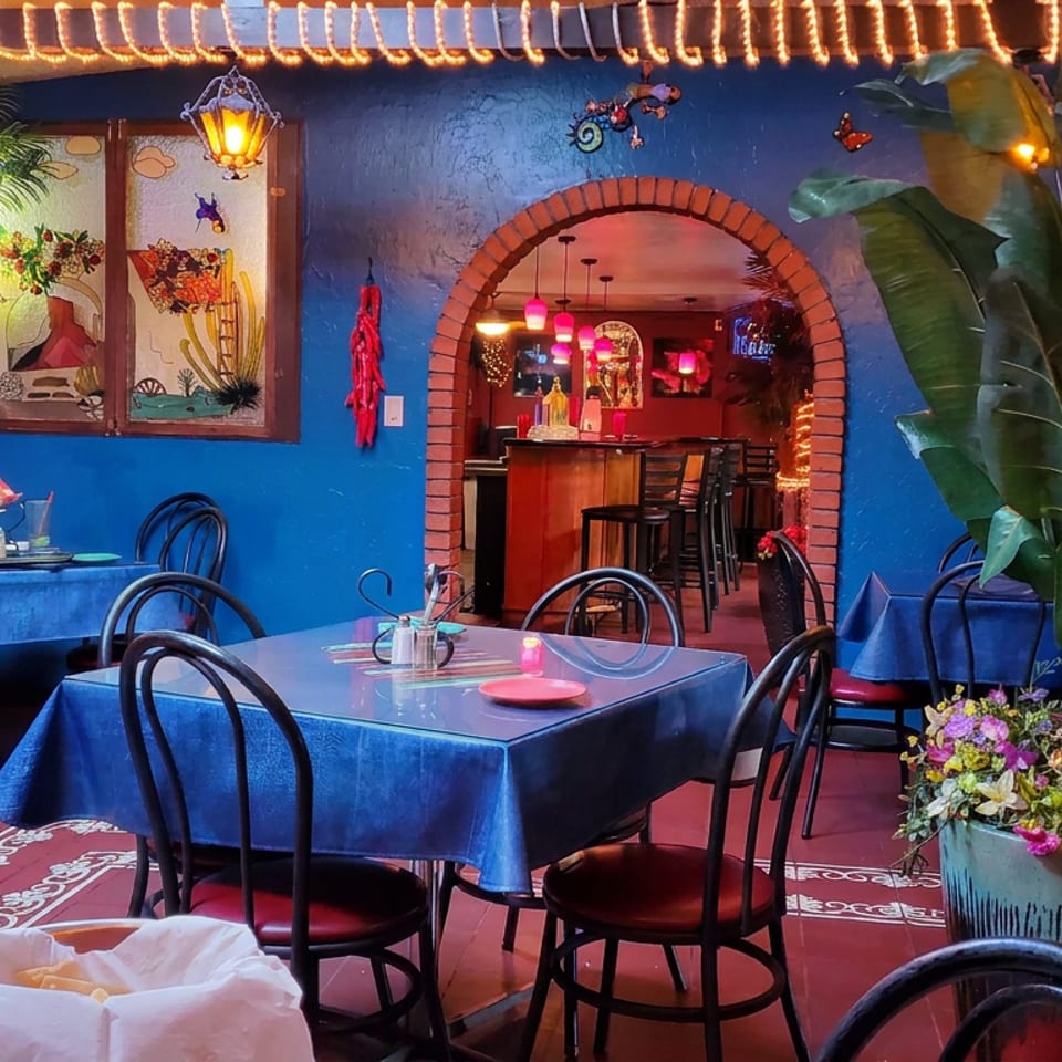 Colorful Mexican-style restaurant dining room with blue tablecloths, string lights, tropical plants, and an arched bar entrance.