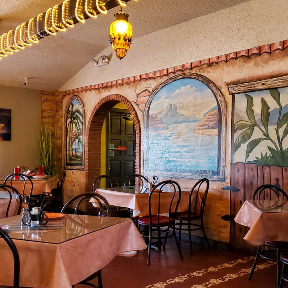 Cozy restaurant dining room with pink tablecloths, black chairs, warm pendant lights, and a mural depicting tropical coastal scenery.