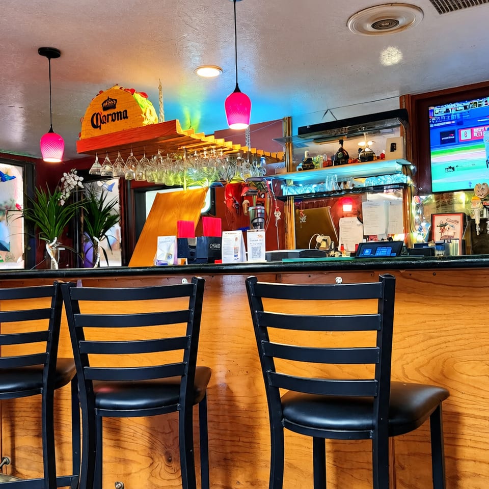 Colorful Mexican restaurant bar with black chairs, hanging glassware, beer taps, sports television, and Corona decorations.
