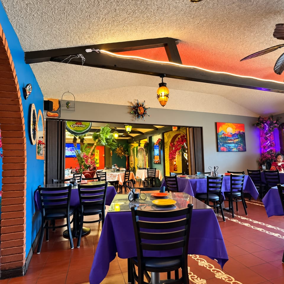 Colorful Mexican restaurant dining room with purple tablecloths, tropical decor, hanging lanterns, and diners.
