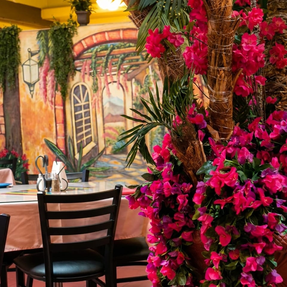 Colorful restaurant interior with black chairs, set tables, tropical plants, and a large arrangement of vivid pink flowers surrounding palm trunks.